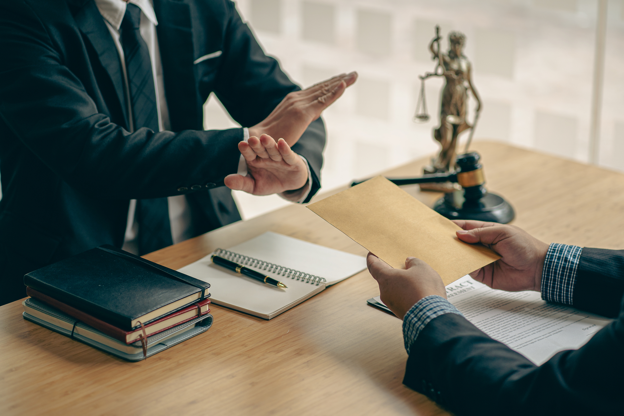 Two individuals in suits are seated at a table; one offers an envelope while the other refuses with a hand gesture, signaling potential legal consequences. Books, a notepad, a gavel, and a small statue of Lady Justice are on the table.
