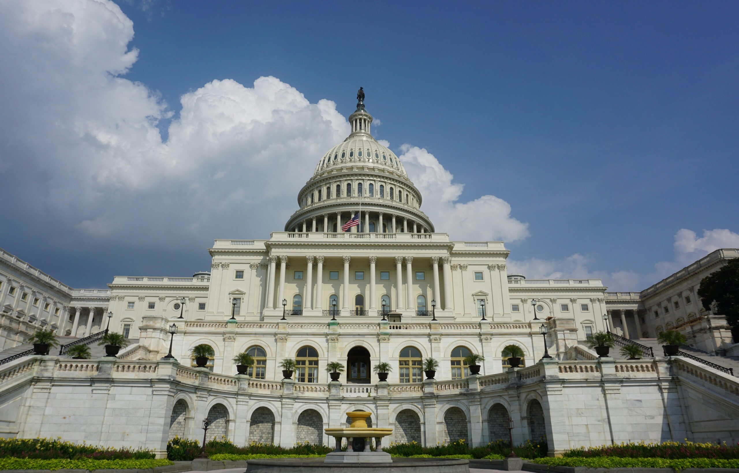 The United States Capitol building beneath a blue sky with clouds, featuring the dome and flag.