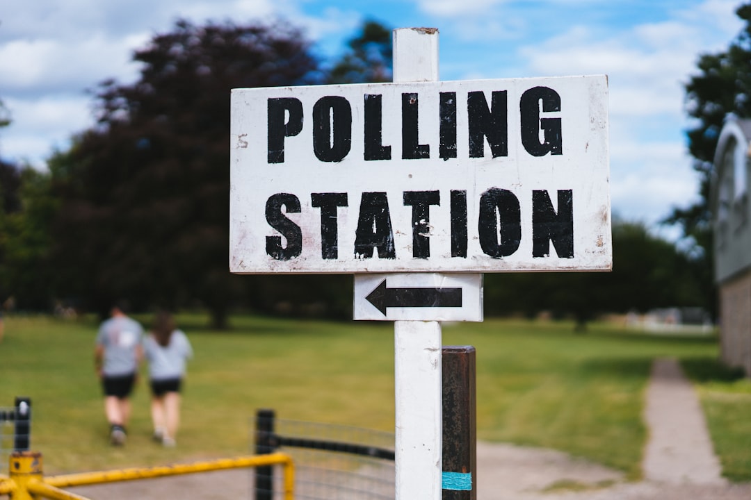 A sign reading "Polling Station" with an arrow pointing left stands in a grassy area, guiding voters on how to elect their leaders. In the background, two people walk purposefully toward their civic duty.