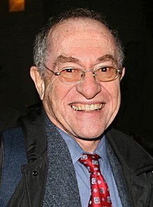 A smiling man wearing glasses, a dark jacket, blue shirt, and red patterned tie.