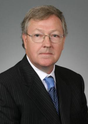 A man in a suit with glasses and a blue tie poses against a gray background.