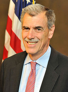 A man in a suit and tie stands in front of an American flag, smiling.