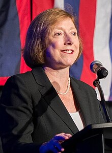 Woman in formal attire speaking at a podium, with red, white, and blue drapes in the background.