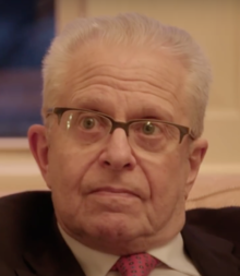 An older man with glasses, wearing a suit and tie, looks slightly to the side.
