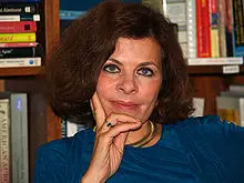 Person with short brown hair resting chin on hand, wearing a blue top, in front of a bookshelf.
