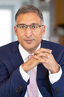 A man wearing glasses and a suit with a pink tie sits with his hands clasped.