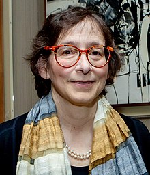 Woman with short brown hair wearing red glasses, a patterned scarf, and a pearl necklace, standing in front of an abstract painting.