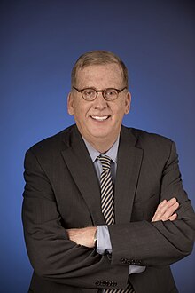Man in a suit with glasses and crossed arms, standing against a blue background.
