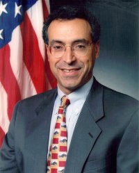 A man in a suit and tie stands in front of a U.S. flag, smiling at the camera.