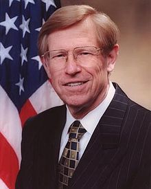 A man with glasses in a suit and tie stands in front of a U.S. flag backdrop.