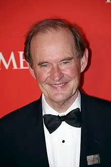 A man wearing a black tuxedo and bow tie stands in front of a red backdrop.
