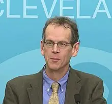 A man in a suit and glasses speaks at a podium against a blue background with partial text visible.