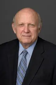 An older man with a bald head, wearing a suit, light blue shirt, and striped tie, poses against a dark background.