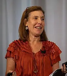 Woman with shoulder-length brown hair wearing a ruffled, rust-colored blouse and glasses on her head, speaking and holding a device, in front of a grey background.
