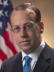 A person in a suit and glasses stands in front of an American flag backdrop.