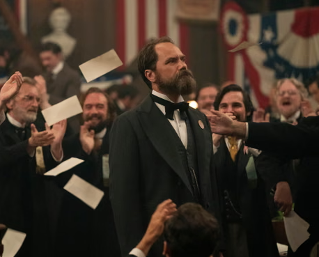A man in formal 19th-century attire stands amid a crowd of applauding men; papers are thrown in the air, and American flags decorate the background.