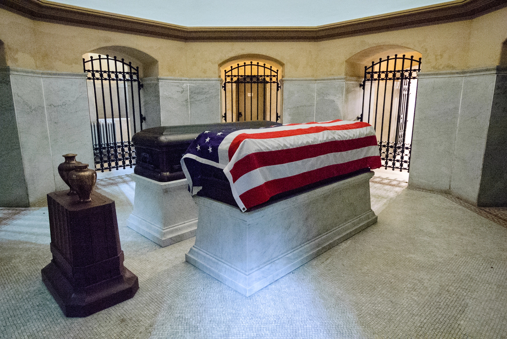 A marble sarcophagus draped with an American flag stands beside a dark coffin and an urn in a gated, round, stone chamber.