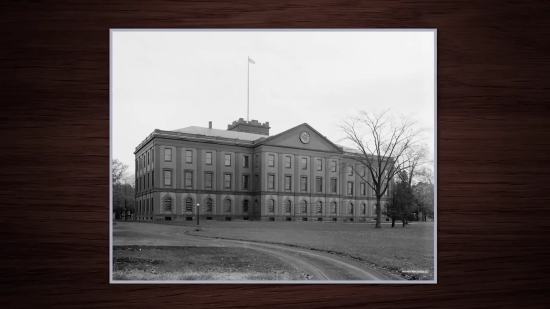 A large, historic brick building with a central clock tower, surrounded by a grassy landscape and bare trees, under an overcast sky.